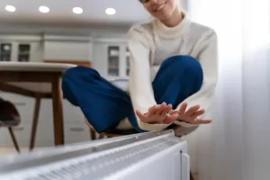 An image of someone warming their hands over a radiator to show the work that can be done to prepare a heating system for winter given that it is now summer.